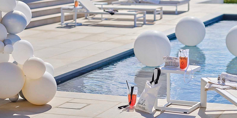 Διακοσμητικό σκηνικό για bachelorette pool party δίπλα στην πισίνα, με άσπρα μπαλόνια, κοκτέιλ και τσάντα με την επιγραφή "Bride to Be".